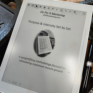 May include: A white e-reader showing a weightlifting methodology titled "Purpose & Intensity Set by Set" with the website GoFitmentoring.com. The text reads "A weightlifting methodology focused on stimulating maximum muscle growth." A black pen is next to the e-reader.