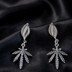May include: A pair of silver-toned drop earrings featuring a small, textured leaf-shaped stud at the top, connected by a jump ring to a larger, detailed seven-pointed leaf charm with prominent veins and a metallic finish.