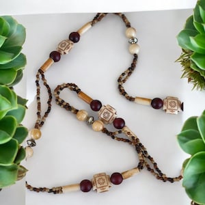 May include: A necklace with a variety of beads in shades of brown, beige, and burgundy. The necklace features small, round beads, larger cylindrical beads, and square beads with carved floral designs. The necklace is displayed on a white surface.
