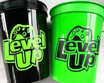 May include: Two plastic cups with a black and green design. The cups have the words "Level Up" and a video game controller graphic.