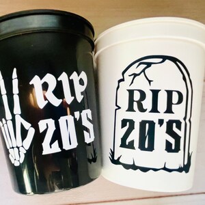Pu&ograve; includere: Due bicchieri di plastica neri e bianchi con il testo "RIP 20's" su di essi. Un bicchiere ha una mano scheletrica che fa il segno della pace e l'altro bicchiere ha una grafica di lapide.