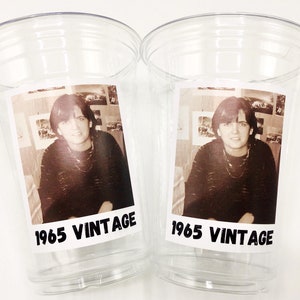 May include: Two clear plastic cups with black and white photos of a person inside. The photos are labelled "1965 Vintage".