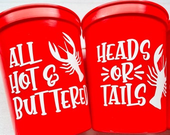 May include: Three red plastic cups with white text and a crawfish design. The cups say "All Hot & Buttered", "Heads or Tails", and "Let's Get Cray".