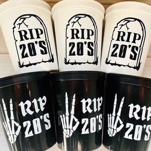 Pu&ograve; includere: Sei bicchieri di plastica, tre bianchi e tre neri, con il testo "RIP 20's" su ciascuno. I bicchieri bianchi hanno un'immagine di una lapide, mentre i bicchieri neri hanno un'immagine di una mano scheletrica.