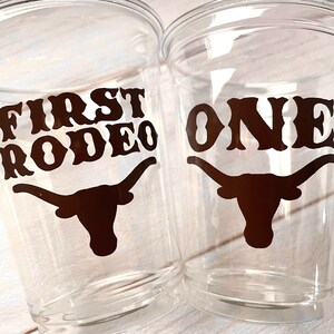 K&ouml;nnte beinhalten: Zwei durchsichtige Plastikbecher mit schwarzen Vinyl-Aufklebern. Ein Becher sagt "First Rodeo" mit einer Silhouette eines Longhorn-Stiers. Der andere Becher sagt "One" mit einer Silhouette eines Longhorn-Stiers.