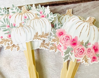 May include: White pumpkin-shaped paper cutouts with gold glitter accents on top and around the edges. Pink flowers and green leaves surround the pumpkins. The cutouts are attached to wooden sticks.