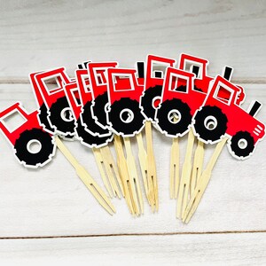May include: A set of red tractor cupcake toppers with black wheels. The toppers are on wooden skewers.