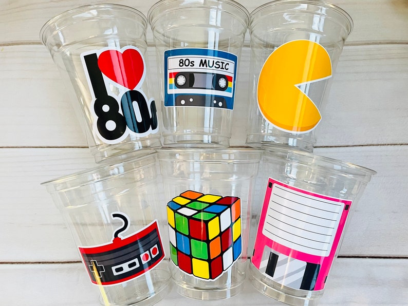 K&ouml;nnte beinhalten: Sechs durchsichtige Plastikbecher mit bunten Aufklebern zum Thema der 80er Jahre. Die Aufkleber zeigen ein Herz mit dem Text "I love 80s", ein Kassettenband mit dem Text "80s Music", einen Pac-Man-Geist, einen Rubik's Cube, einen Joystick und eine Diskette.