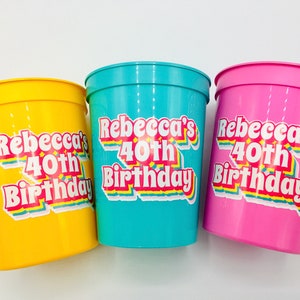 Puede incluir: Tres vasos de pl&aacute;stico en amarillo, turquesa y rosa. Cada vaso tiene una pegatina que dice "Rebecca's 40th Birthday" en una fuente retro con acentos de arco&iacute;ris. Los vasos son probablemente para una fiesta de cumplea&ntilde;os.