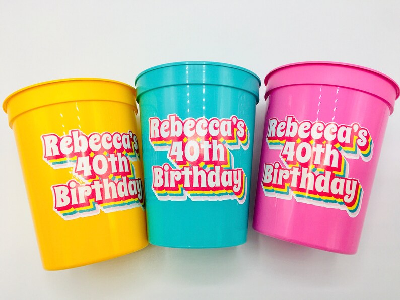 Puede incluir: Tres vasos de pl&aacute;stico en amarillo, turquesa y rosa. Cada vaso tiene una pegatina que dice "Rebecca's 40th Birthday" en una fuente retro con acentos de arco&iacute;ris. Los vasos son probablemente para una fiesta de cumplea&ntilde;os.