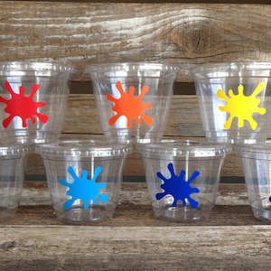 Peut inclure: Six gobelets en plastique transparent avec des motifs de peinture &eacute;clabouss&eacute;s de couleurs. Les gobelets sont dispos&eacute;s en rang&eacute;e sur une surface en bois. Les couleurs sont le rouge, l'orange, le jaune, le vert, le bleu et le violet.