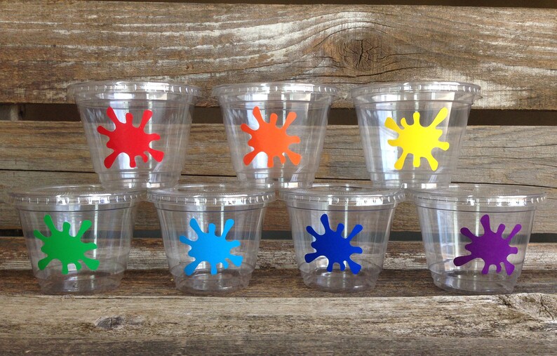 Peut inclure: Six gobelets en plastique transparent avec des motifs de peinture &eacute;clabouss&eacute;s de couleurs. Les gobelets sont dispos&eacute;s en rang&eacute;e sur une surface en bois. Les couleurs sont le rouge, l'orange, le jaune, le vert, le bleu et le violet.