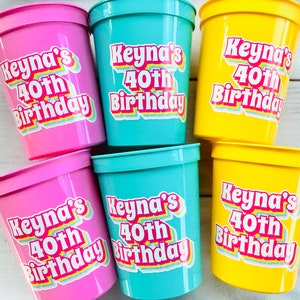 Puede incluir: Un conjunto de vasos de pl&aacute;stico rosas, turquesas y amarillos. Cada vaso tiene una pegatina con la frase "Keyna's 40th Birthday" en una fuente retro colorida. Los vasos son probablemente para una fiesta de cumplea&ntilde;os.