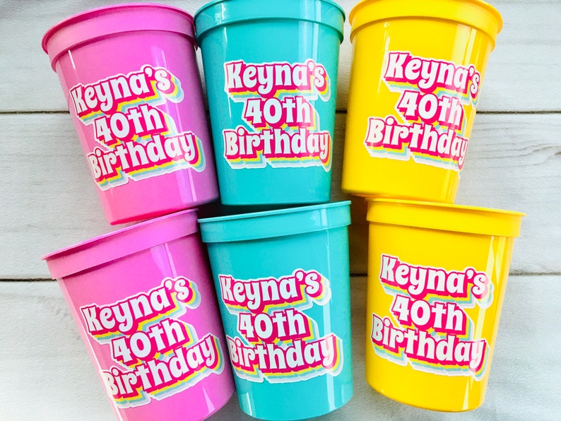Puede incluir: Un conjunto de vasos de pl&aacute;stico rosas, turquesas y amarillos. Cada vaso tiene una pegatina con la frase "Keyna's 40th Birthday" en una fuente retro colorida. Los vasos son probablemente para una fiesta de cumplea&ntilde;os.