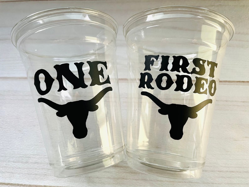 K&ouml;nnte beinhalten: Zwei durchsichtige Plastikbecher mit schwarzen Vinyl-Aufklebern. Ein Becher sagt "ONE" und der andere sagt "FIRST RODEO". Beide Becher zeigen eine schwarze Silhouette eines Longhorn-Stiers.