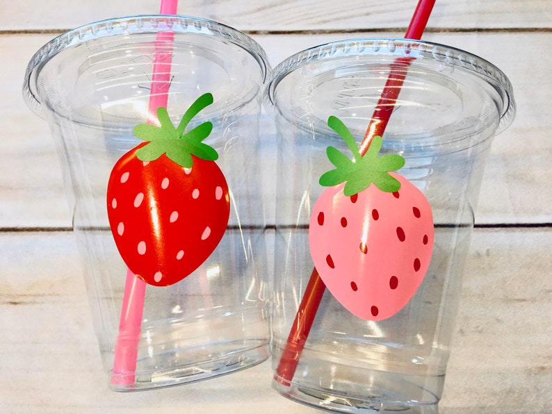 Puede incluir: Dos vasos de pl&aacute;stico transparentes con pajitas y dise&ntilde;os de fresas. Un vaso tiene una fresa roja y una pajita rosa, el otro tiene una fresa rosa y una pajita roja.
