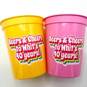 Puede incluir: Dos vasos de fiesta de pl&aacute;stico, uno amarillo y otro rosa, con el texto "Beers & Cheers to Whit's 40 years!" en una pegatina colorida. Los vasos son cil&iacute;ndricos con un borde ligeramente ensanchado.