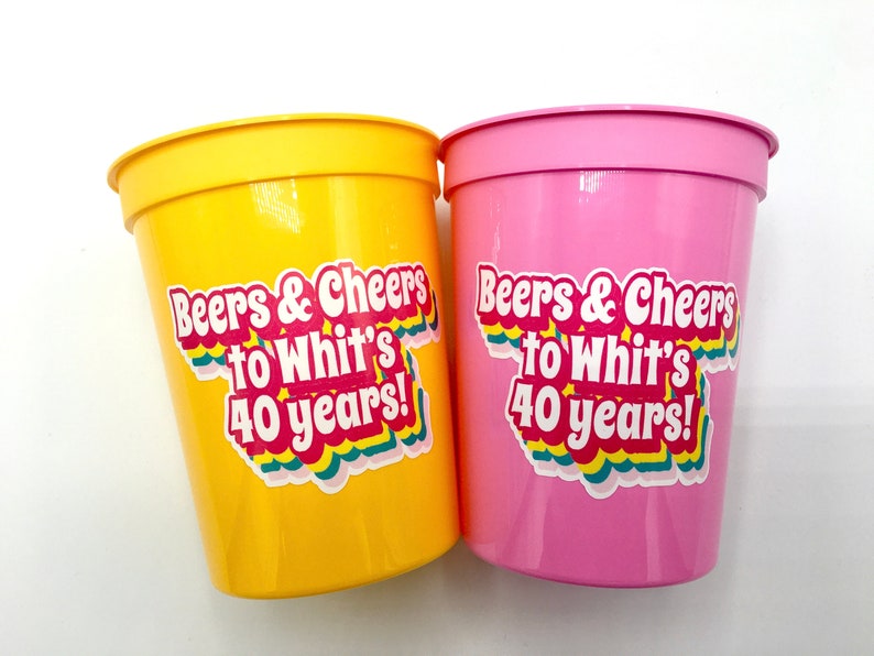 Puede incluir: Dos vasos de fiesta de pl&aacute;stico, uno amarillo y otro rosa, con el texto "Beers & Cheers to Whit's 40 years!" en una pegatina colorida. Los vasos son cil&iacute;ndricos con un borde ligeramente ensanchado.