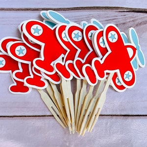 May include: A set of red and blue airplane cupcake toppers with wooden sticks. The airplanes have white stars on them.