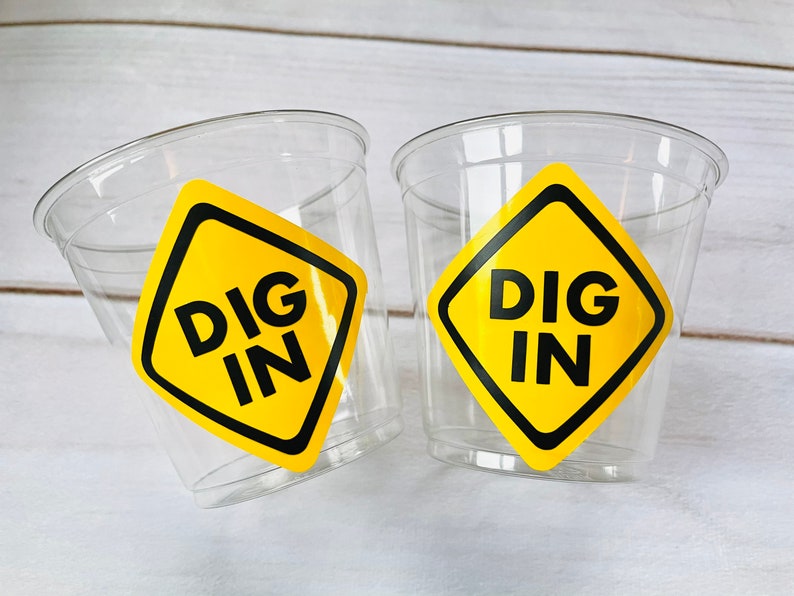 May include: Two clear plastic cups with yellow construction sign stickers that say "DIG IN" in black letters.