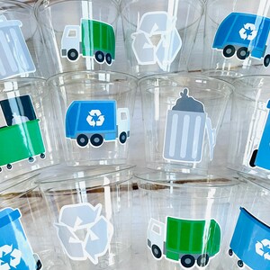 May include: A set of clear plastic cups with various recycling themed stickers. The stickers feature a green recycling bin, a blue recycling bin, a gray recycling bin, a green garbage truck, a blue garbage truck, and a recycle symbol.