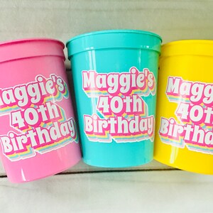 Puede incluir: Tres vasos de fiesta de pl&aacute;stico en rosa, turquesa y amarillo. Cada vaso tiene una pegatina que dice "40 cumplea&ntilde;os de Maggie" en una fuente retro colorida. Los vasos son probablemente para una celebraci&oacute;n de cumplea&ntilde;os.