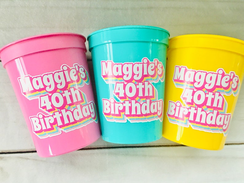 Puede incluir: Tres vasos de fiesta de pl&aacute;stico en rosa, turquesa y amarillo. Cada vaso tiene una pegatina que dice "40 cumplea&ntilde;os de Maggie" en una fuente retro colorida. Los vasos son probablemente para una celebraci&oacute;n de cumplea&ntilde;os.