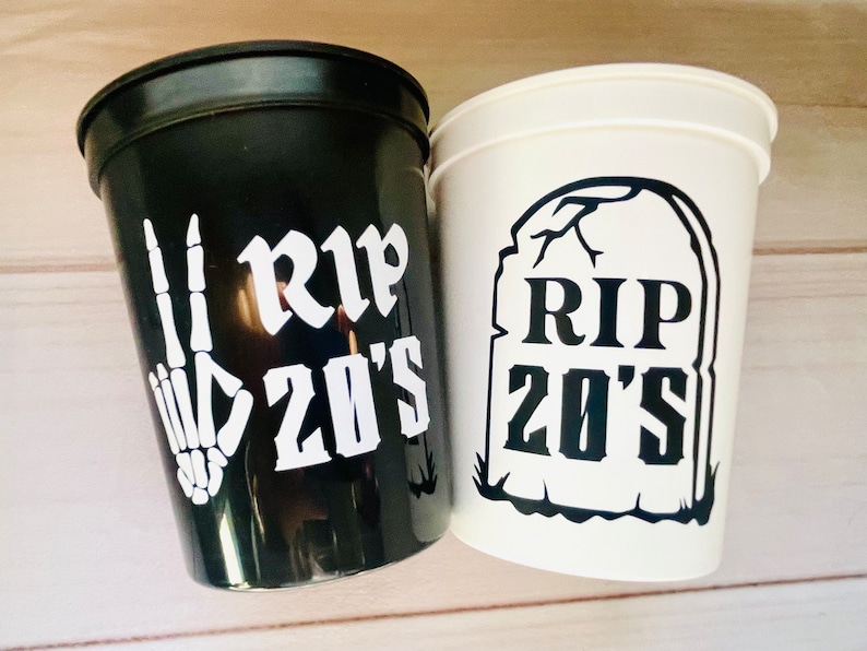 Pu&ograve; includere: Due bicchieri di plastica neri e bianchi con il testo "RIP 20's" su di essi. Un bicchiere ha una mano scheletrica che fa il segno della pace e l'altro bicchiere ha una grafica di lapide.