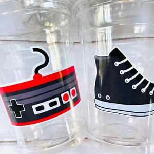 K&ouml;nnte beinhalten: Zwei durchsichtige Plastikbecher mit bunten Aufklebern. Ein Becher hat einen Aufkleber mit einem Retro-Videospielcontroller mit roten und grauen Kn&ouml;pfen. Der andere Becher hat einen Aufkleber mit einem schwarzen High-Top-Sneaker.
