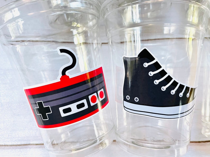 K&ouml;nnte beinhalten: Zwei durchsichtige Plastikbecher mit bunten Aufklebern. Ein Becher hat einen Aufkleber mit einem Retro-Videospielcontroller mit roten und grauen Kn&ouml;pfen. Der andere Becher hat einen Aufkleber mit einem schwarzen High-Top-Sneaker.