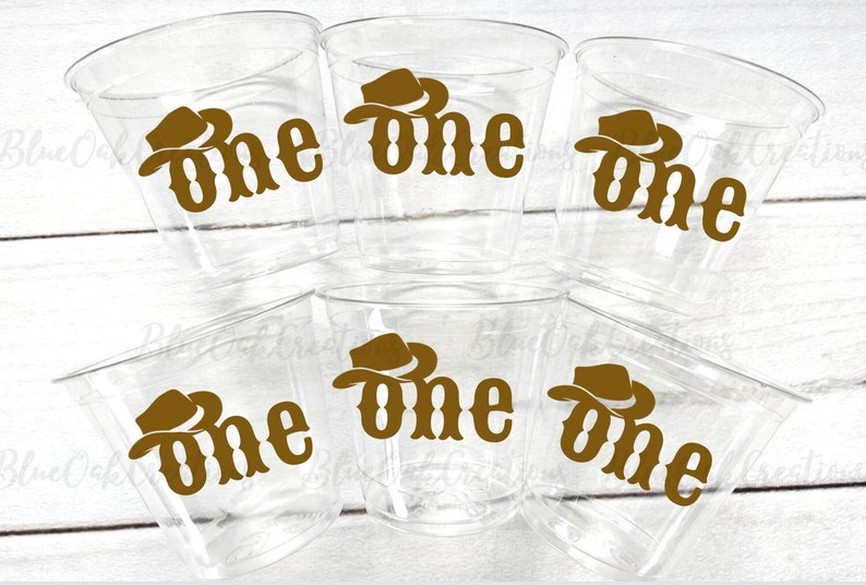 Op de afbeelding: Zes doorzichtige plastic bekers met een bruine cowboyhoed en "one" erop gedrukt.
