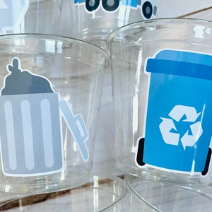May include: Clear plastic cups with blue and gray recycling themed stickers. The stickers feature a trash can with a lid, a recycling bin with a recycle symbol, and a garbage truck.