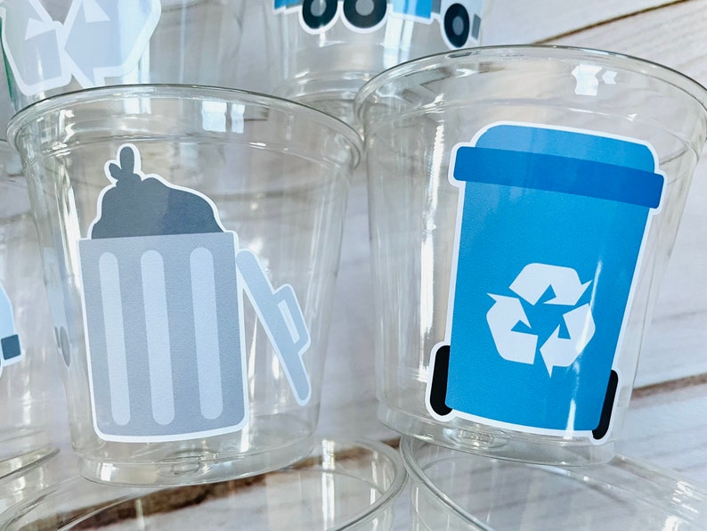 May include: Clear plastic cups with blue and gray recycling themed stickers. The stickers feature a trash can with a lid, a recycling bin with a recycle symbol, and a garbage truck.