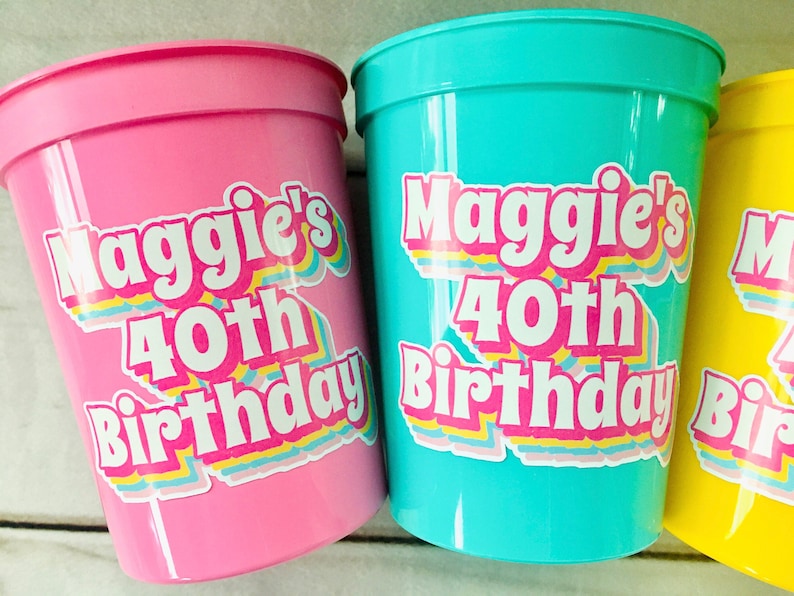 Puede incluir: Tres vasos de pl&aacute;stico en rosa, turquesa y amarillo. Cada vaso tiene una pegatina que dice "40 cumplea&ntilde;os de Maggie" en una fuente retro colorida. Los vasos son probablemente para una fiesta de cumplea&ntilde;os.
