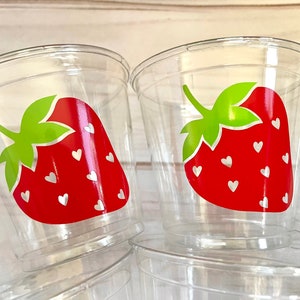 Op de afbeelding: Doorzichtige plastic bekers met een rood aardbeienontwerp. De aardbeien hebben witte hartjes en groene bladeren.