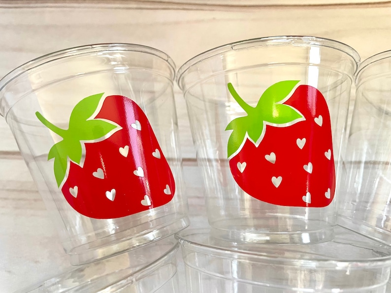 Op de afbeelding: Doorzichtige plastic bekers met een rood aardbeienontwerp. De aardbeien hebben witte hartjes en groene bladeren.