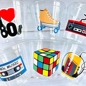 May include: Six clear plastic cups with colorful 80s themed stickers. The stickers feature a heart with "I love 80s", a pair of roller skates, a video game controller, a Rubik's Cube, a cassette tape with "80s Music", and a cassette tape with headphones.