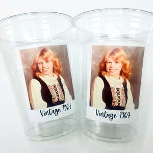 May include: Two clear plastic cups featuring a vintage photograph of a woman with blonde hair, wearing a black and white patterned shirt. The photo is labelled "Vintage 1964" on each cup.