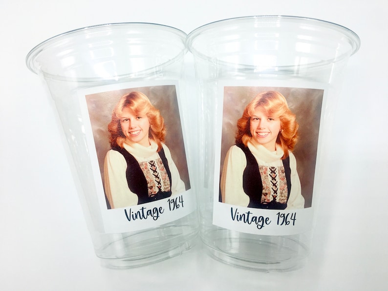 May include: Two clear plastic cups featuring a vintage photograph of a woman with blonde hair, wearing a black and white patterned shirt. The photo is labelled "Vintage 1964" on each cup.