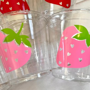 Op de afbeelding: Transparante plastic bekers met een roze aardbeidesontwerp en witte hartjes. De aardbeien hebben groene bladeren.