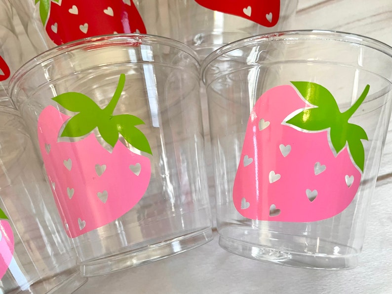 Op de afbeelding: Transparante plastic bekers met een roze aardbeidesontwerp en witte hartjes. De aardbeien hebben groene bladeren.