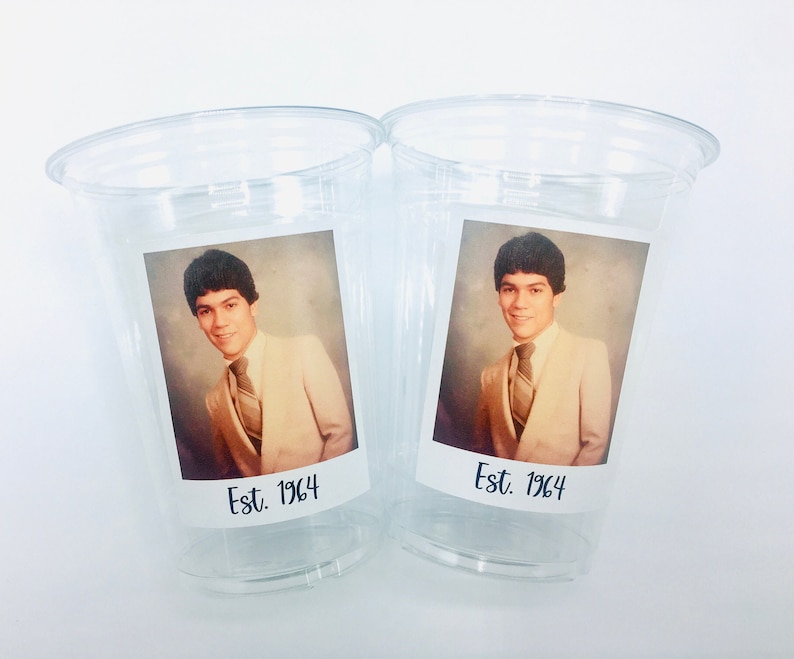 May include: Two clear plastic cups with a photo of a person in a suit and tie. The photo is in a white frame with the text "Est. 1964" below it.