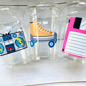 K&ouml;nnte beinhalten: Drei durchsichtige Plastikbecher mit Designs im 80er-Jahre-Stil. Der erste Becher hat einen blauen und grauen Ghettoblaster mit gelben Blitzen. Der zweite Becher hat einen gelben und blauen Rollschuh. Der dritte Becher hat eine rosa Diskette mit einem wei&szlig;en Etikett.