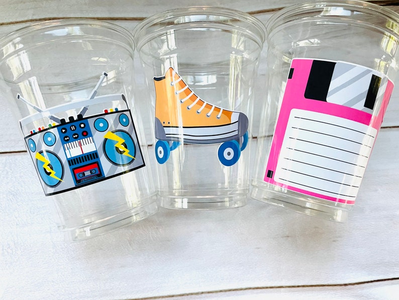 K&ouml;nnte beinhalten: Drei durchsichtige Plastikbecher mit Designs im 80er-Jahre-Stil. Der erste Becher hat einen blauen und grauen Ghettoblaster mit gelben Blitzen. Der zweite Becher hat einen gelben und blauen Rollschuh. Der dritte Becher hat eine rosa Diskette mit einem wei&szlig;en Etikett.