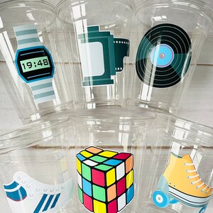 K&ouml;nnte beinhalten: Sechs durchsichtige Plastikbecher mit bunten Aufklebern im 80er-Jahre-Stil. Die Aufkleber zeigen eine Digitaluhr, eine Filmrolle, eine Schallplatte, einen Rubik's Cube, ein Paar Rollschuhe und ein Paar Turnschuhe.