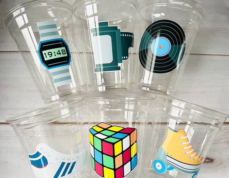 K&ouml;nnte beinhalten: Sechs durchsichtige Plastikbecher mit bunten Aufklebern im 80er-Jahre-Stil. Die Aufkleber zeigen eine Digitaluhr, eine Filmrolle, eine Schallplatte, einen Rubik's Cube, ein Paar Rollschuhe und ein Paar Turnschuhe.