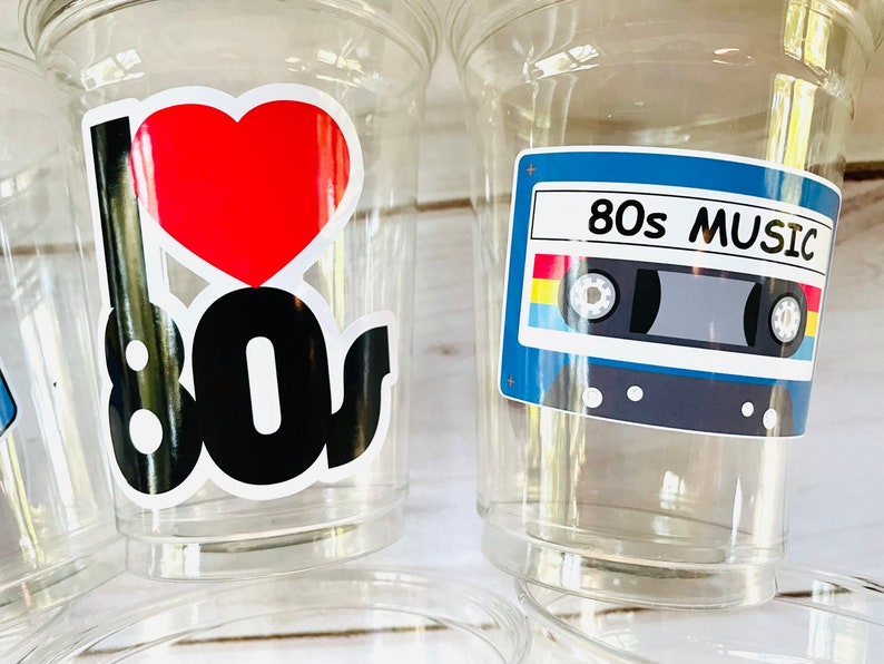 K&ouml;nnte beinhalten: Zwei durchsichtige Plastikbecher mit Designs im 80er-Jahre-Stil. Ein Becher hat ein rotes Herz und den Text "I 80s". Der andere Becher hat ein blaues Kassettenband mit dem Text "80s MUSIC".