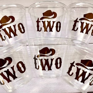 Op de afbeelding: Zes doorzichtige plastic bekers met een bruine cowboyhoed en "TWO" erop gedrukt. De bekers zijn op elkaar gestapeld.