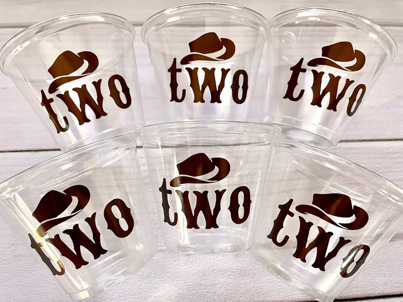 Op de afbeelding: Zes doorzichtige plastic bekers met een bruine cowboyhoed en "TWO" erop gedrukt. De bekers zijn op elkaar gestapeld.