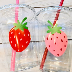 Puede incluir: Dos vasos de pl&aacute;stico transparentes con pajitas y dise&ntilde;os de fresas. Un vaso tiene una fresa roja y una pajita rosa, el otro tiene una fresa rosa y una pajita roja.